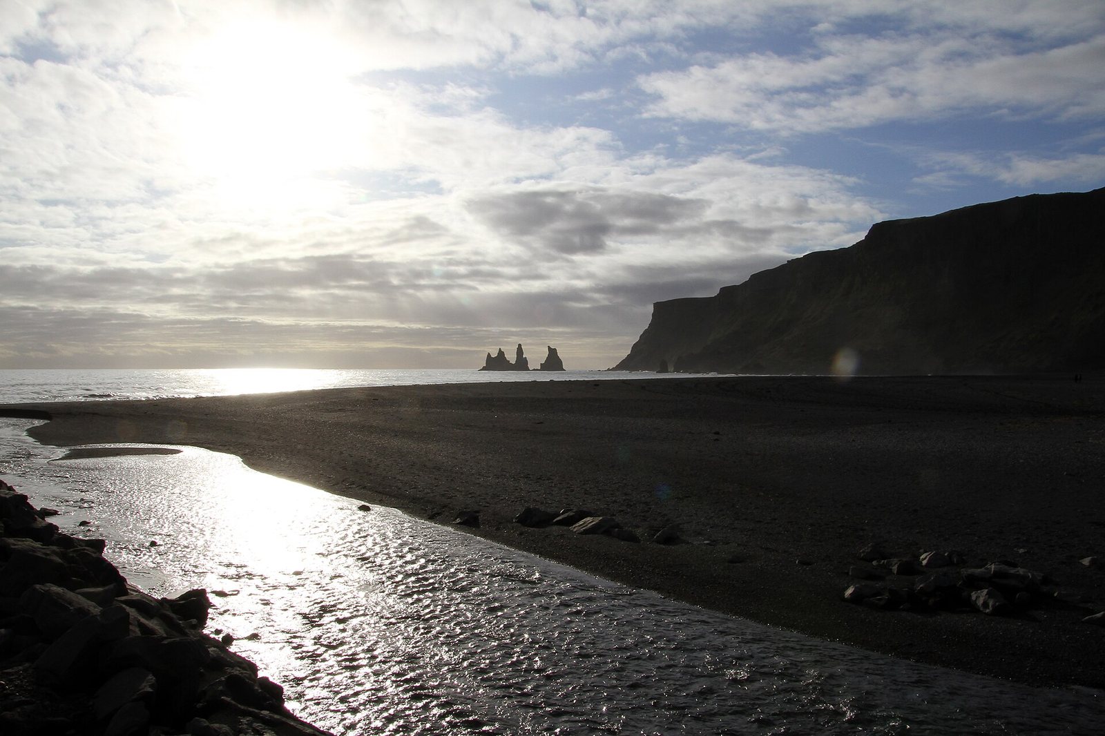 A black sand beach with a stream reflecting sunlight in the foreground, the ocean to the left, and two distant sea stacks against a dark cliff under a cloudy sky.