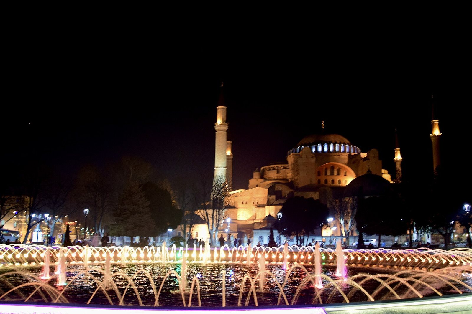 The Hagia Sophia Grand Mosque is illuminated at night, seen from across a large fountain with multiple coloured water jets in the foreground. Trees and streetlights are also visible.