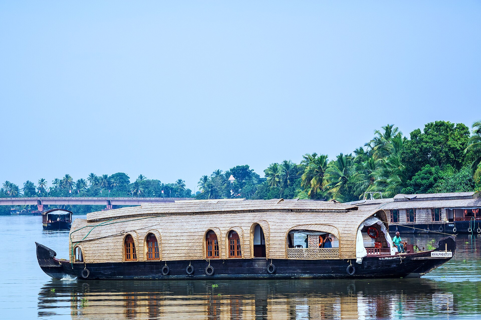 A kettuvallam houseboat with an arched cane roof gliding on still green backwater between palm-lined banks.