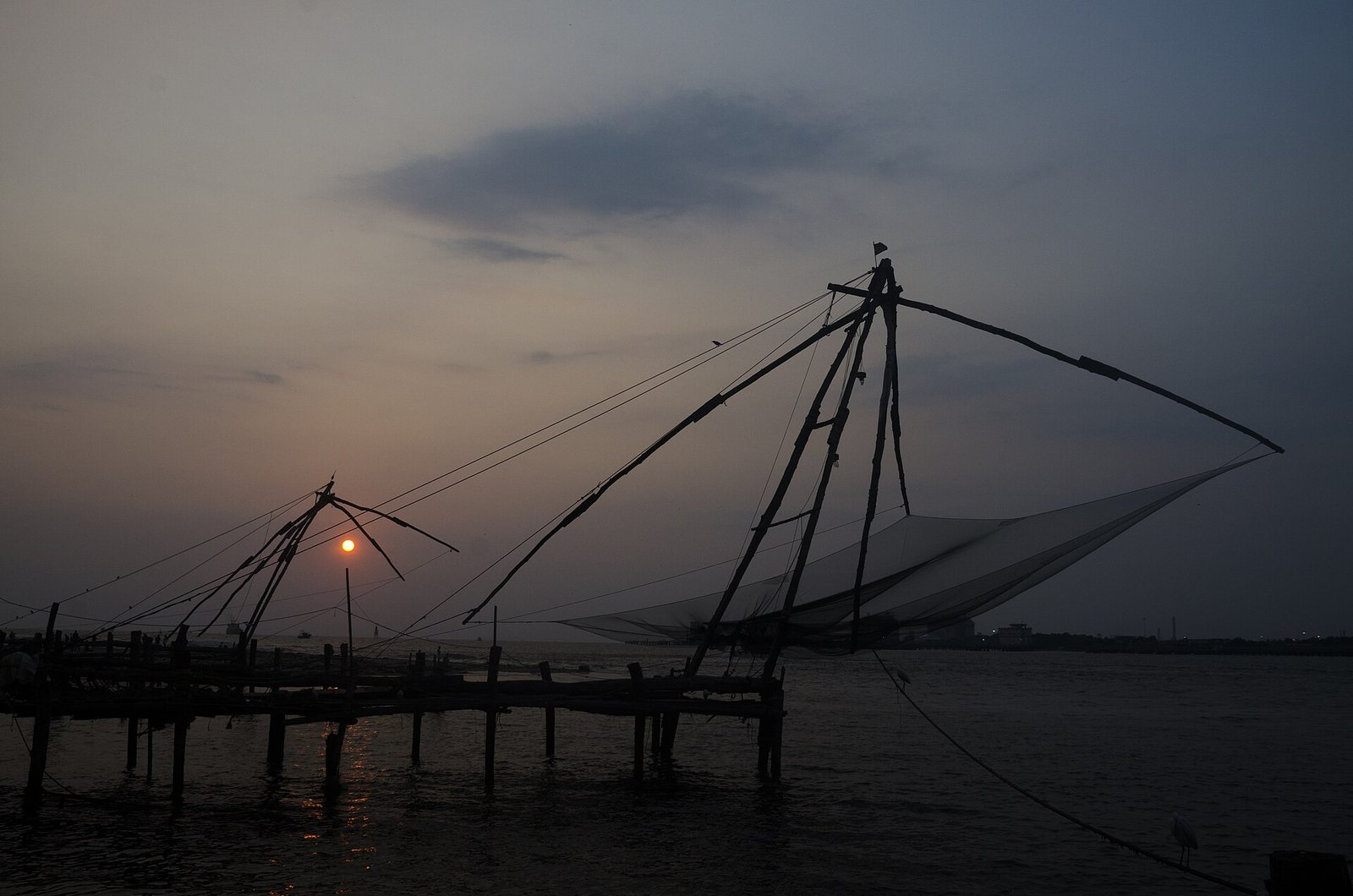 A sunset over water, with the sun visible between the dark silhouettes of two Chinese fishing nets at Fort Kochi.