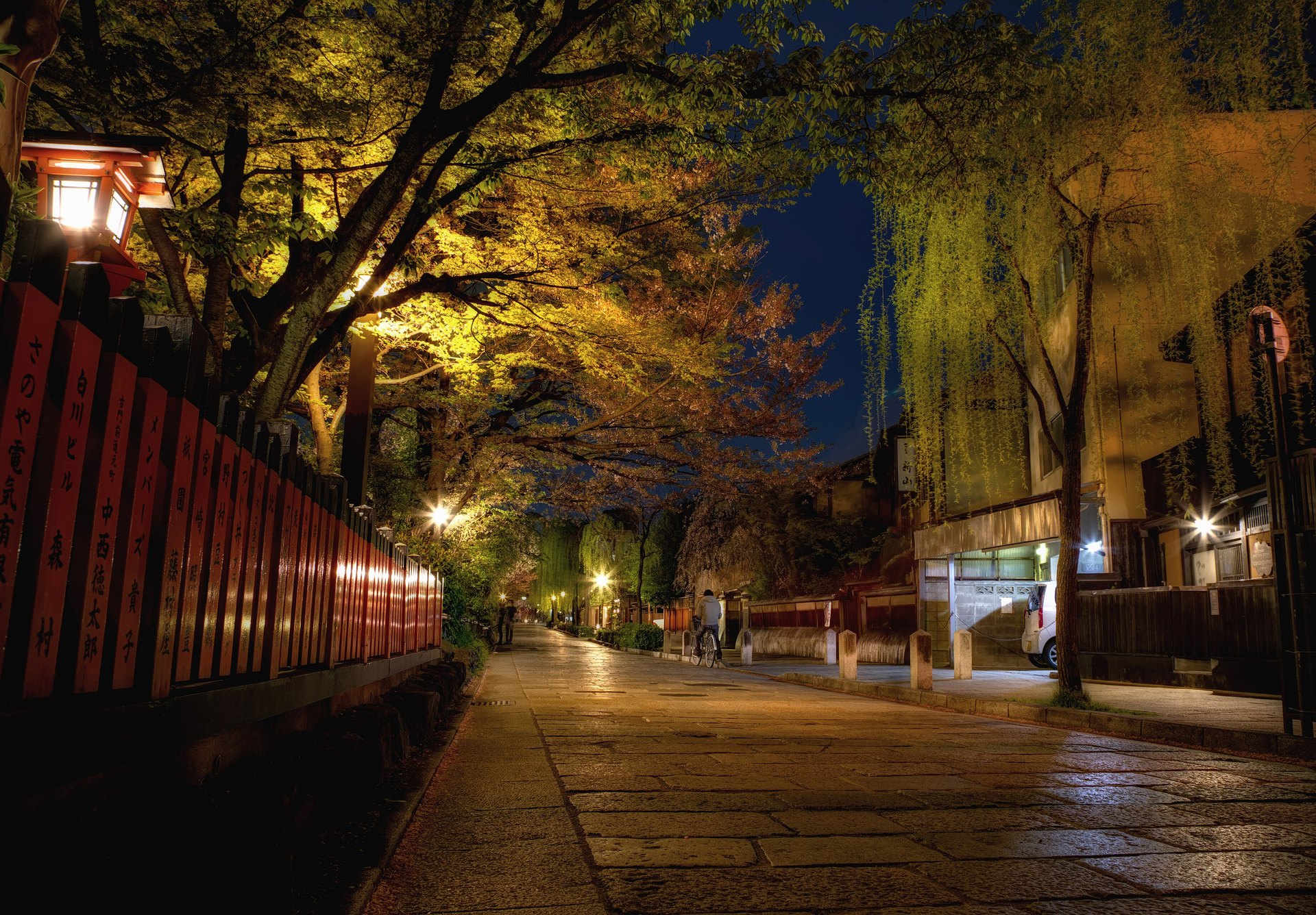 A stone-paved lane in Gion at night, lit by lanterns under trees, with a red lacquered fence on the left and wooden buildings on the right