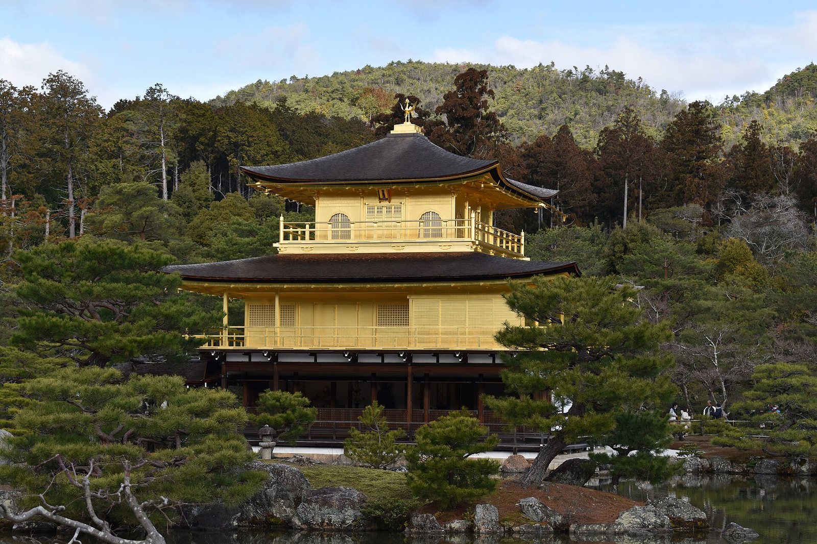 A three-story golden pavilion with dark roofs stands by a pond, surrounded by green and brown trees, with a forested hill in the background.