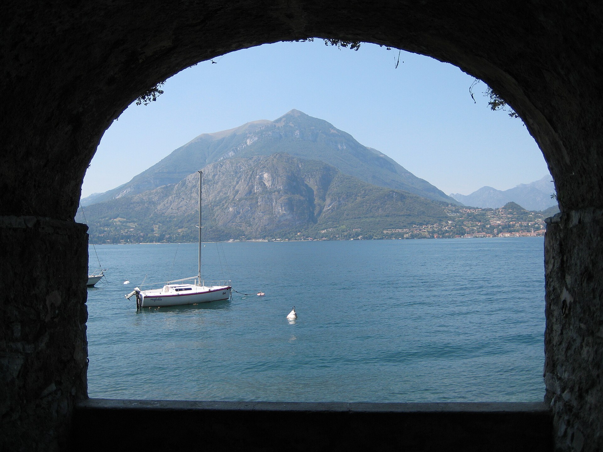 The lakefront promenade at Menaggio on the western shore of Lake Como, with a tree-lined walkway, moored boats, and mountains across the water.