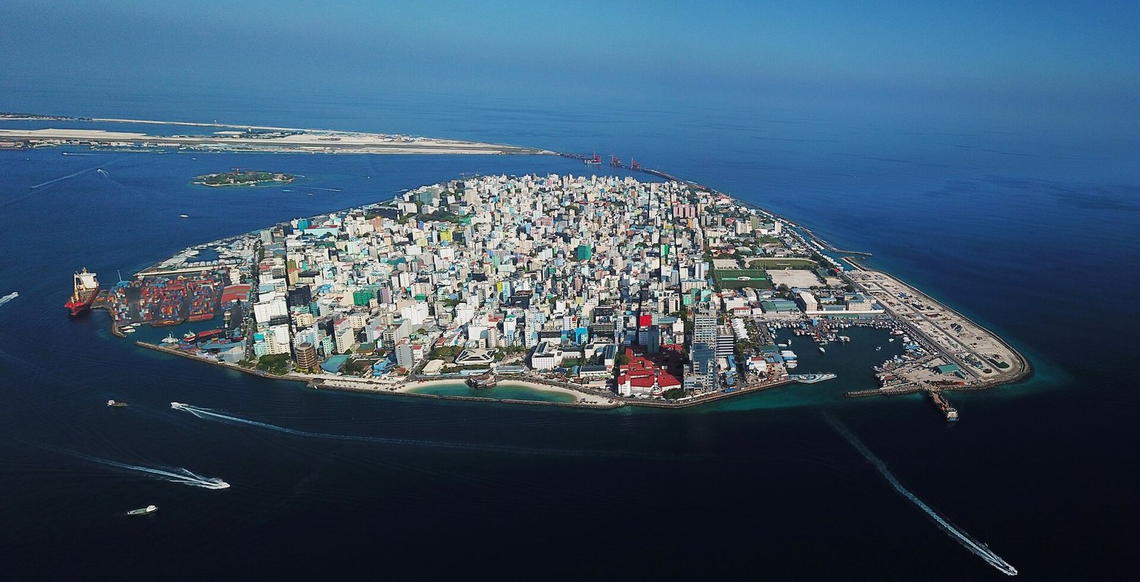 An aerial view shows a densely built-up island city surrounded by blue ocean, with a busy harbour, several boats, and a distant landmass.