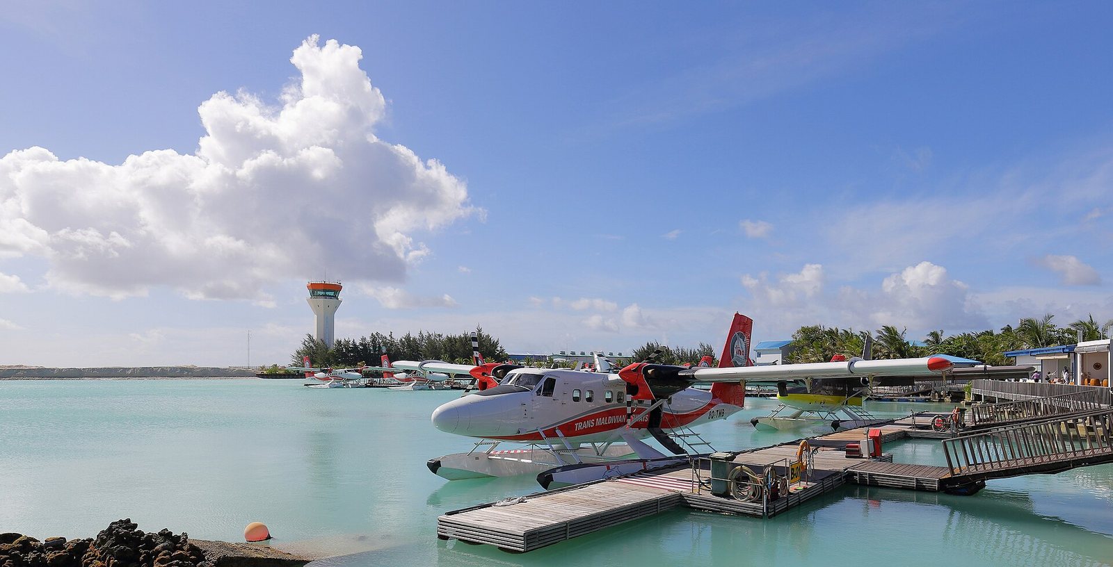 A Trans Maldivian Airways DHC-6 Twin Otter seaplane is docked at a pier over turquoise water, with other seaplanes and an airport control tower in the background.