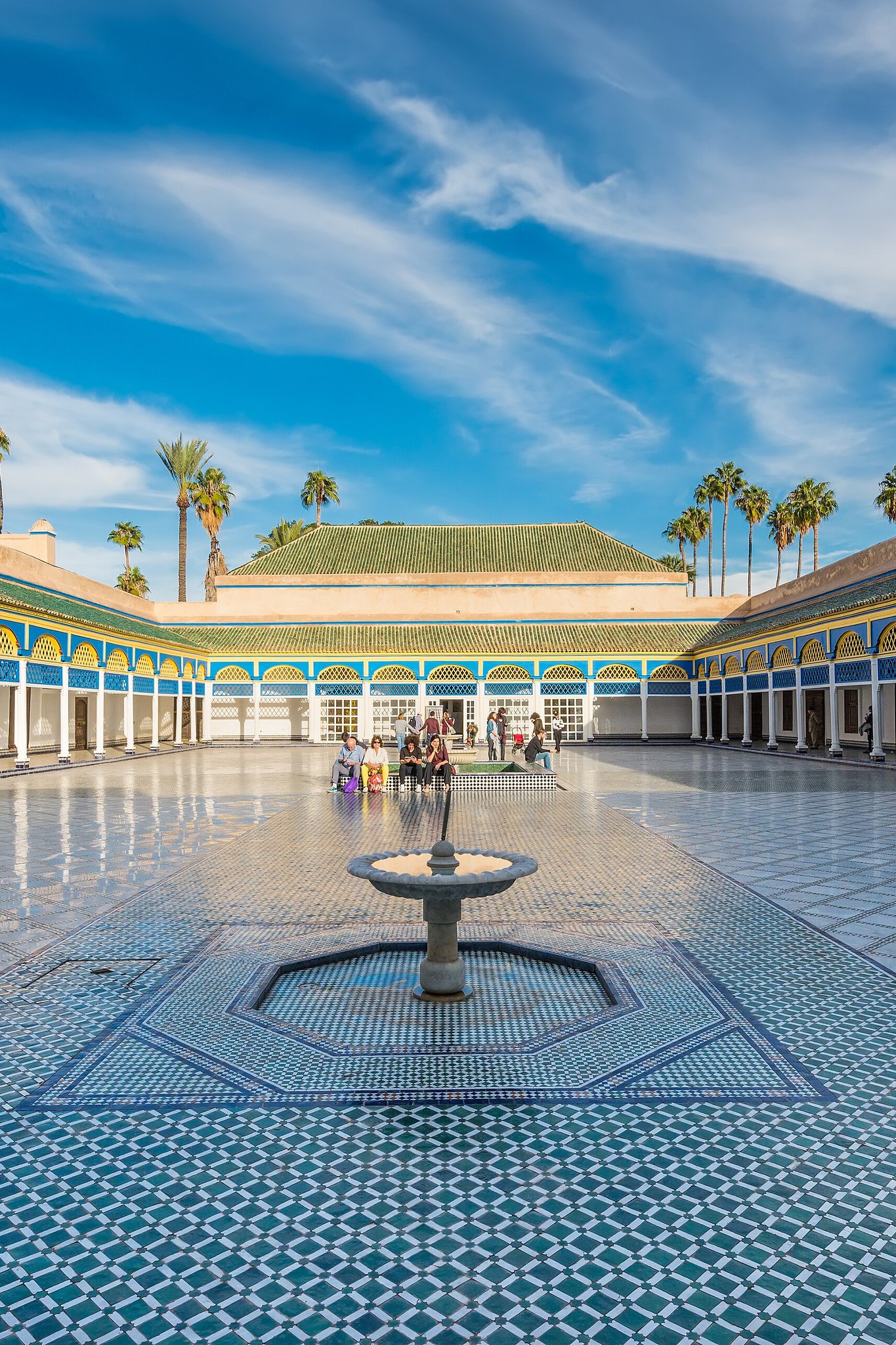 A courtyard paved in blue and white tile with a central fountain, surrounded by arcades with painted ceilings under a bright blue sky