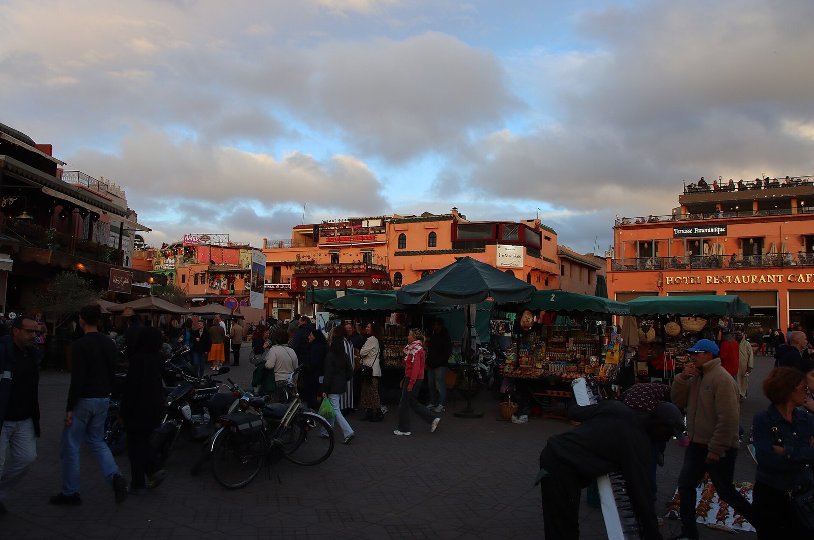 A busy market square at dusk with crowds, canvas-topped stalls and orange buildings with cafe terraces around it