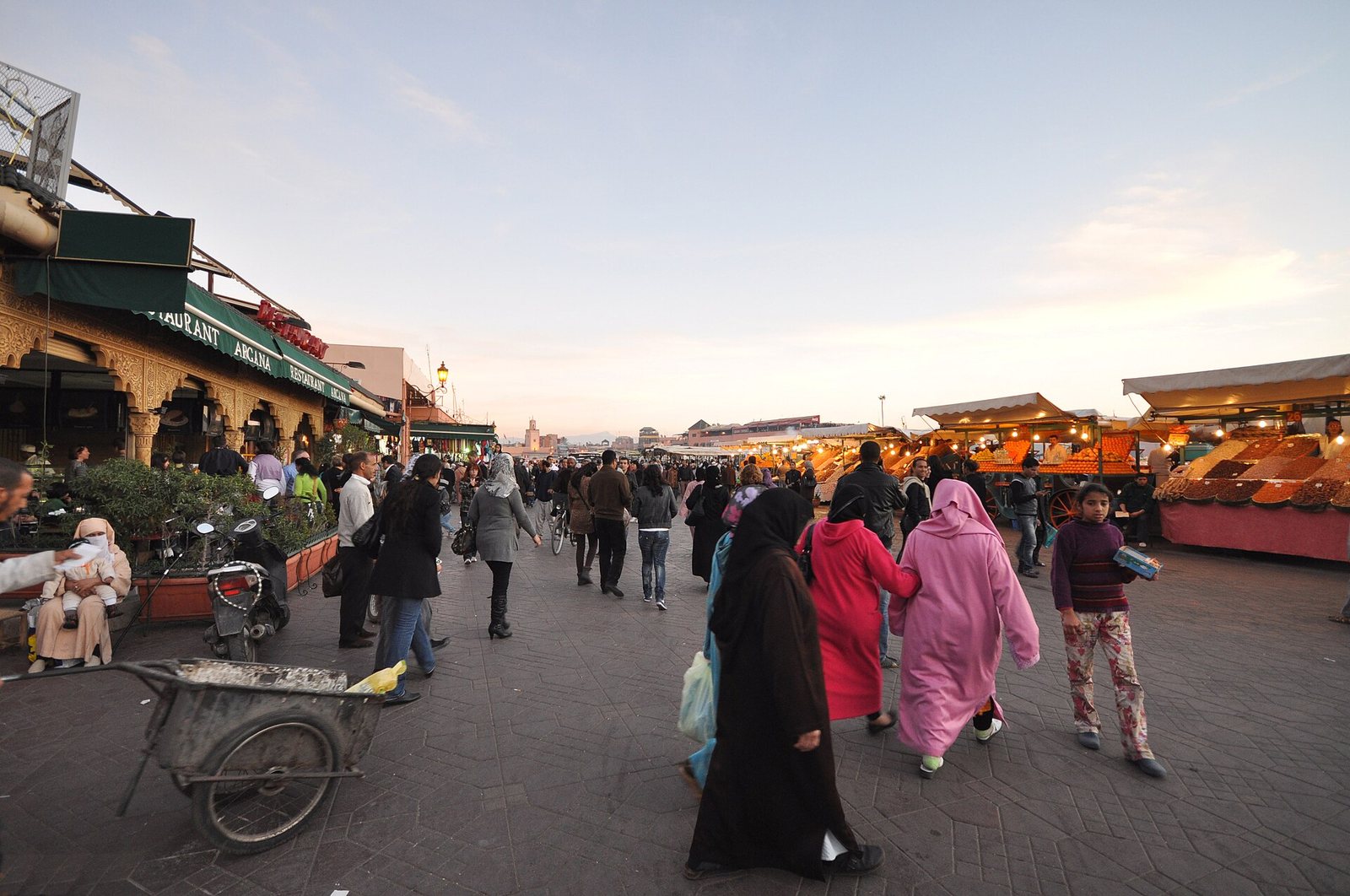A busy market square at dusk with many people walking, illuminated stalls on the right, and buildings with awnings on the left. A cart is in the foreground.