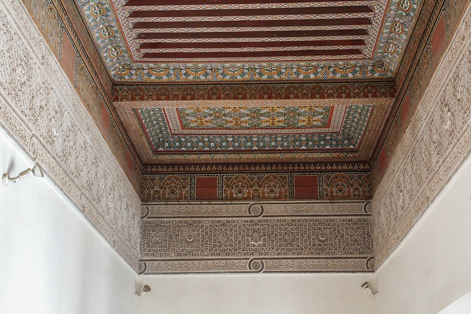A room corner shows a painted wooden ceiling with geometric and floral patterns, and carved plasterwork on the upper walls above plain white lower walls.