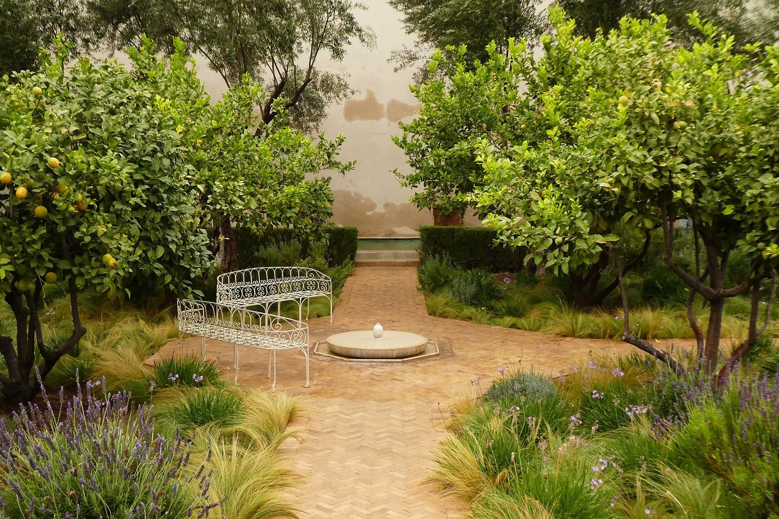 A garden with a cross-shaped brick path, a central fountain, two white benches, green trees with yellow fruit, and various plants, with a light-coloured wall in the background.