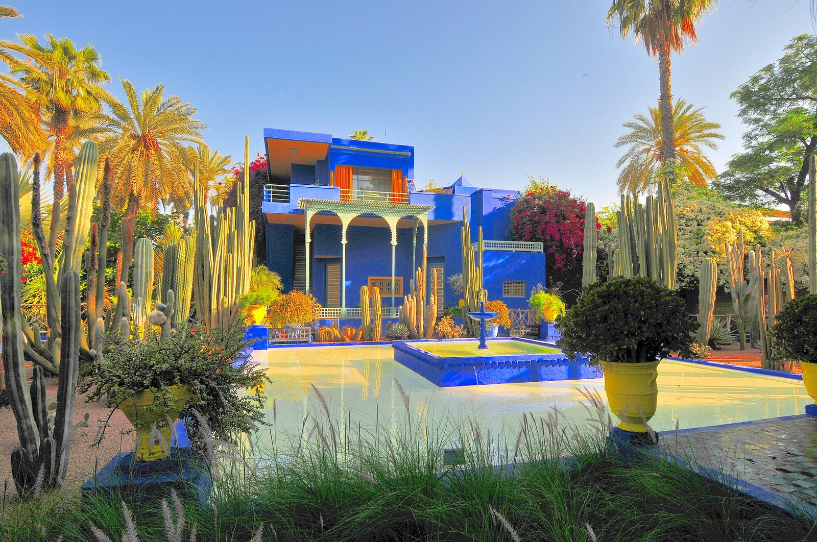 A blue building with orange trim stands amidst tall cacti and palm trees, with a rectangular pool and fountain in the foreground under a clear sky.