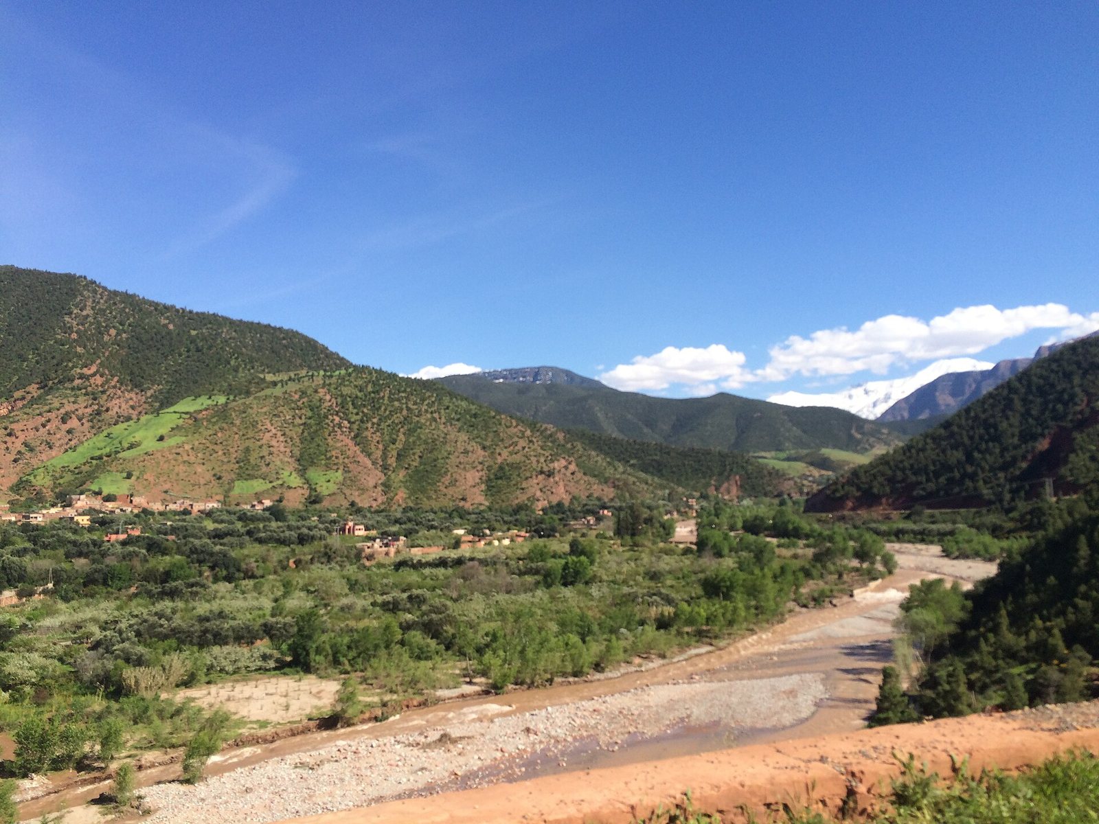 A wide landscape view shows a riverbed flowing through a green valley with small villages, surrounded by mountains, some with snow, under a blue sky.