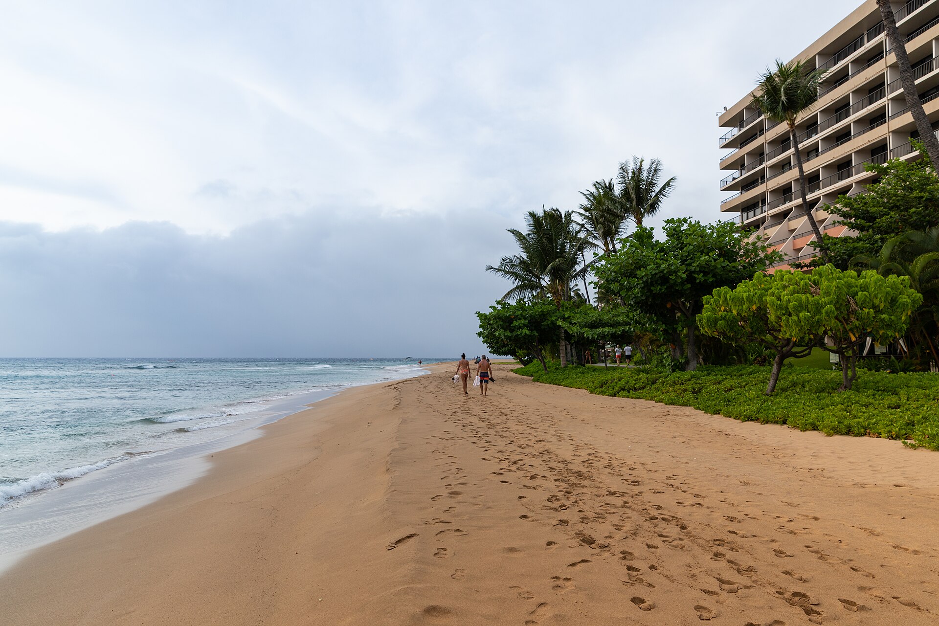 Kāʻanapali Beach on the west coast of Maui seen from the air, a long curve of pale sand backed by resorts and palms, with the black lava headland of Puʻu Kekaʻa at the far end and calm blue water offshore.
