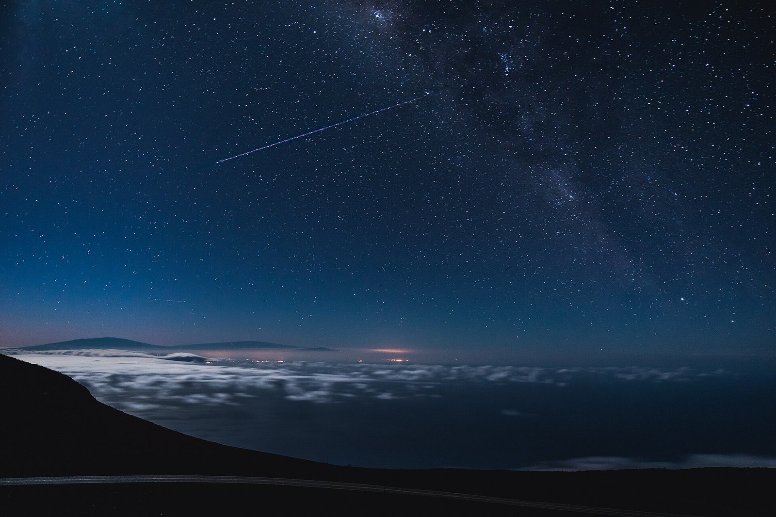 A night sky shows numerous stars and a light trail above a dark landscape with a curving road, a layer of clouds, and distant land with faint lights.