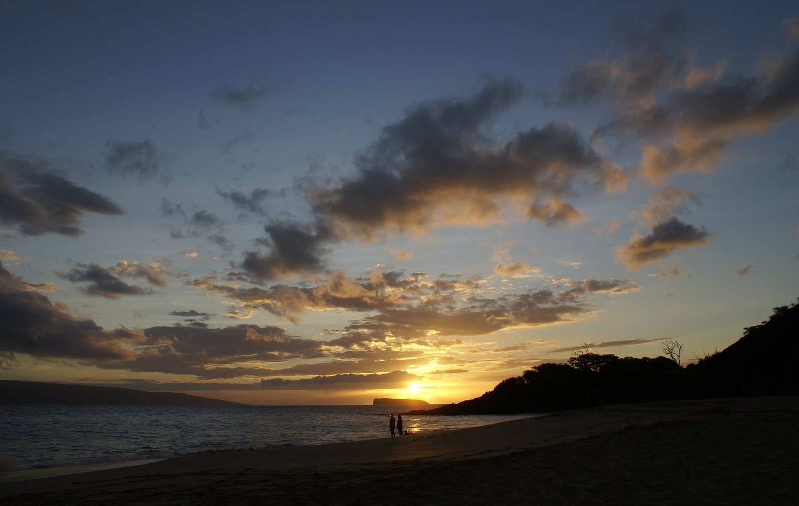 A sunset over a beach shows two silhouetted figures by the water, a distant landmass, and a hillside under a sky with clouds.