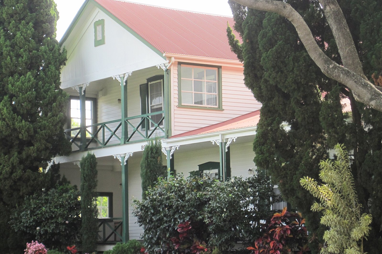A white two-storey wooden house with a red roof, green-railed balconies and a front porch among trees and shrubs
