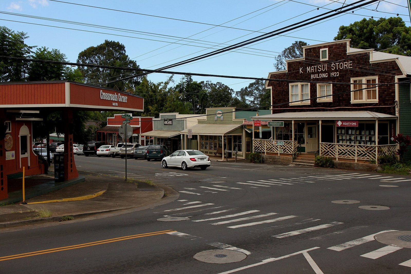A street intersection in a town with several buildings, including 'K Matsui Store Building 1923' and 'Crossroads Corner', with parked and moving cars.