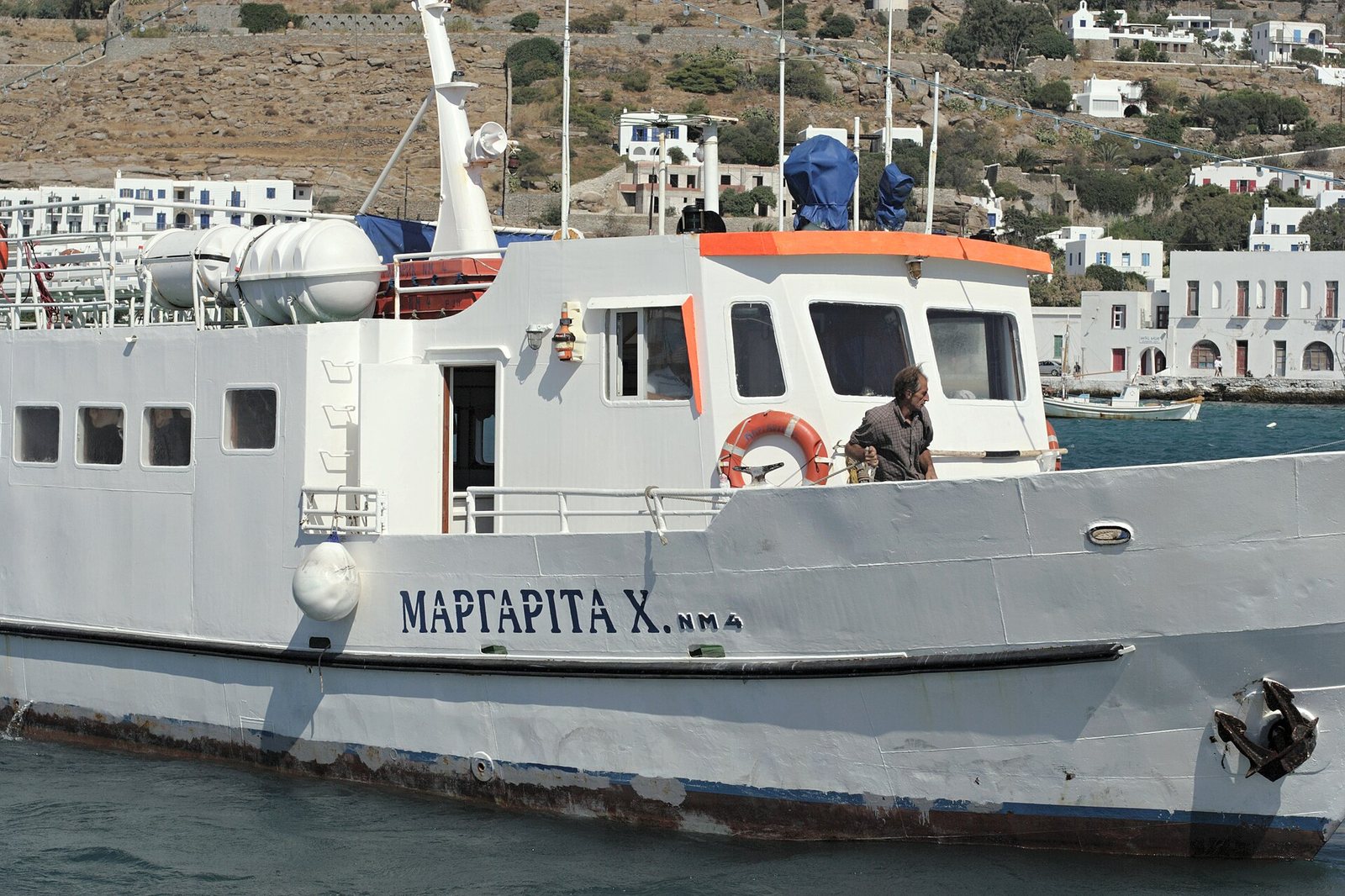 A white ship named Margarita with a man on deck is in the foreground, with white buildings on a hillside in the background.