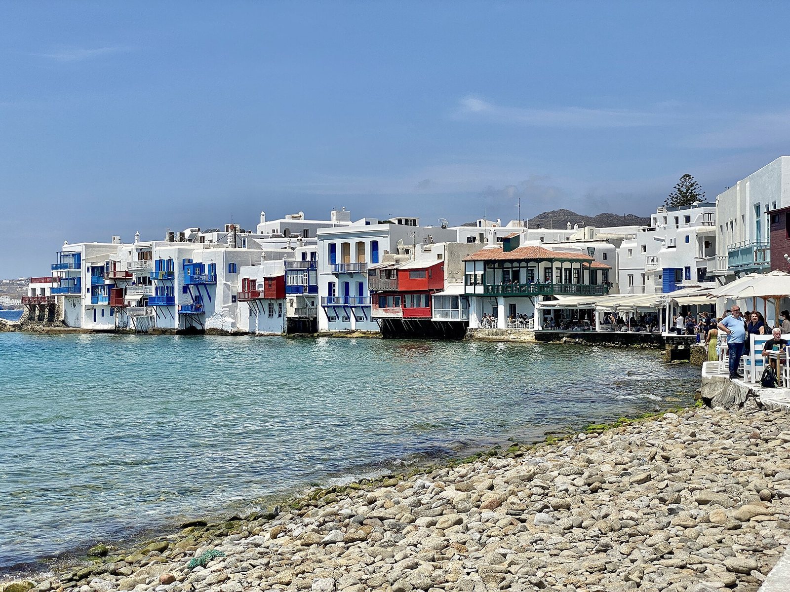 White houses with blue, red, and green balconies line the clear blue sea, with a rocky shore in the foreground and outdoor cafes on the right under a blue sky.