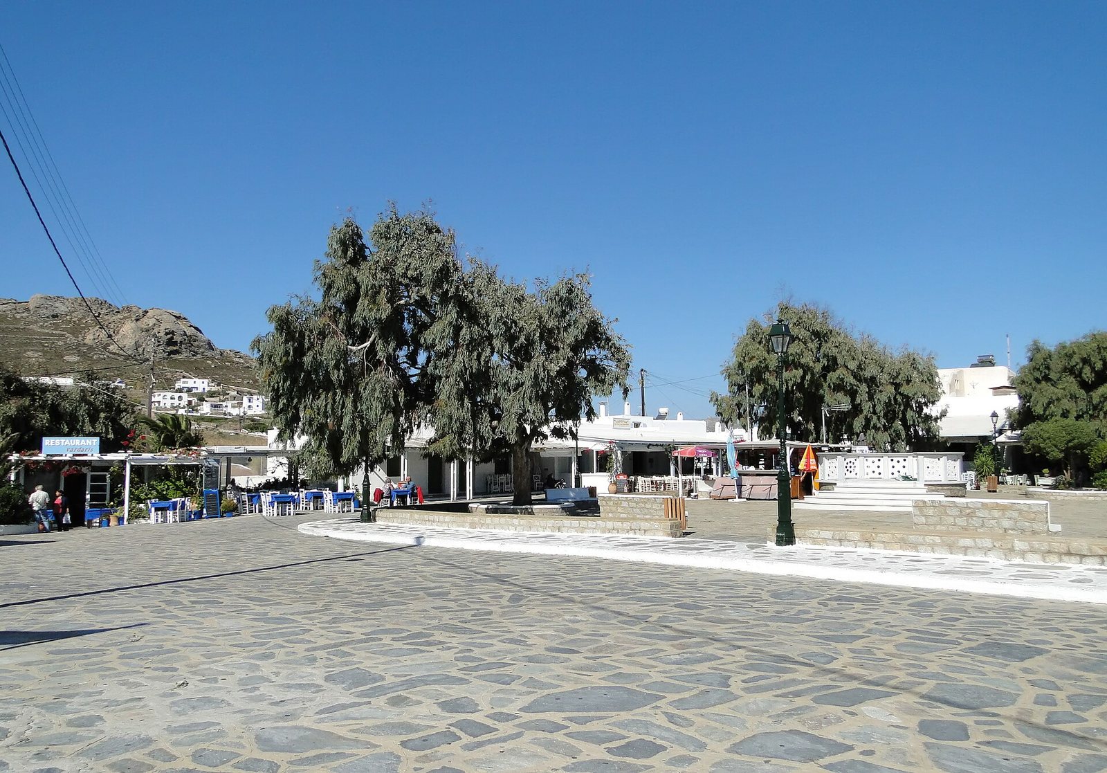A paved village square with large trees, white buildings housing restaurants with outdoor seating, and a rocky hill with more white houses in the background under a clear sky.