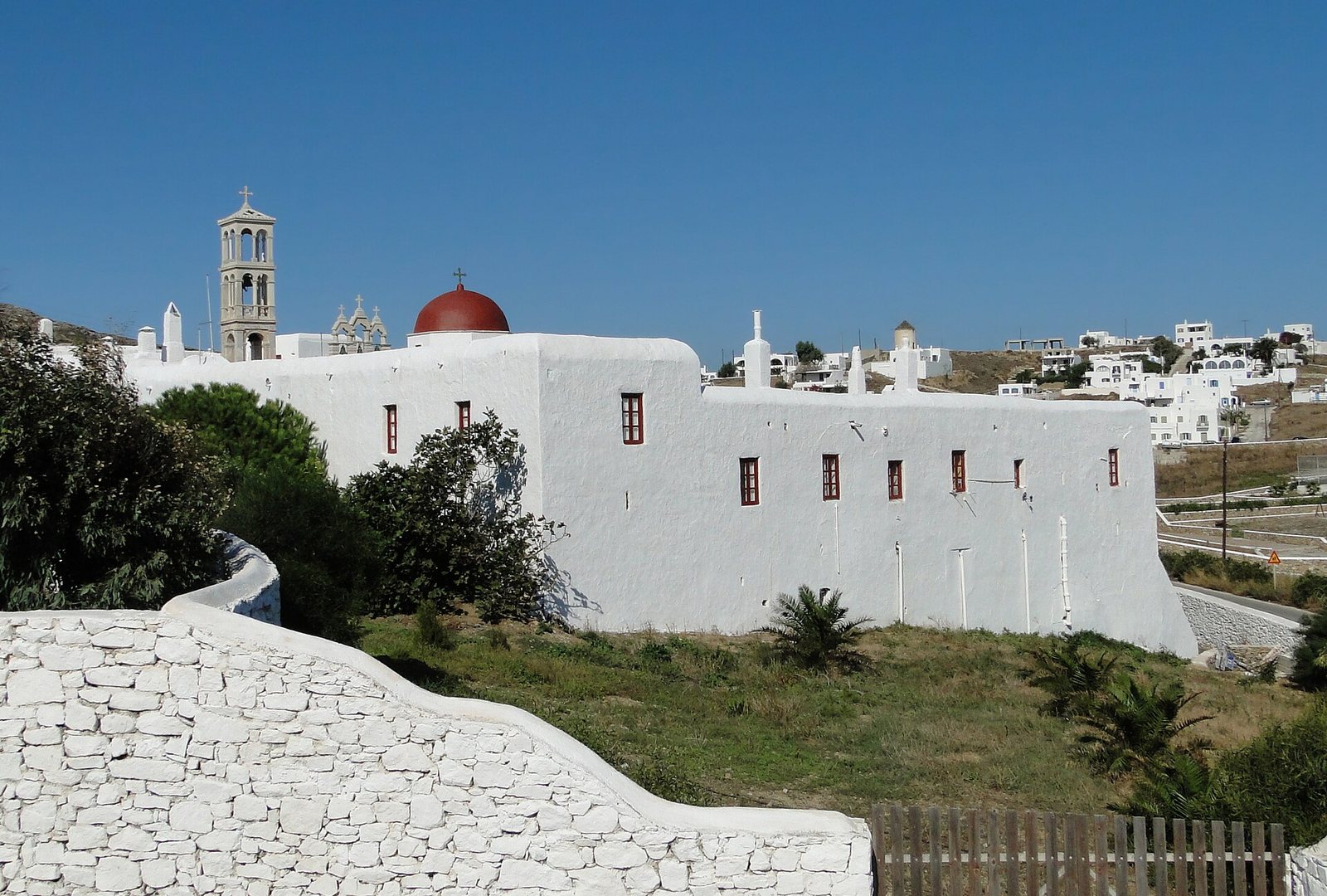 A white-washed monastery building with a red dome and a bell tower stands on a grassy slope under a clear blue sky, with white village buildings in the background.