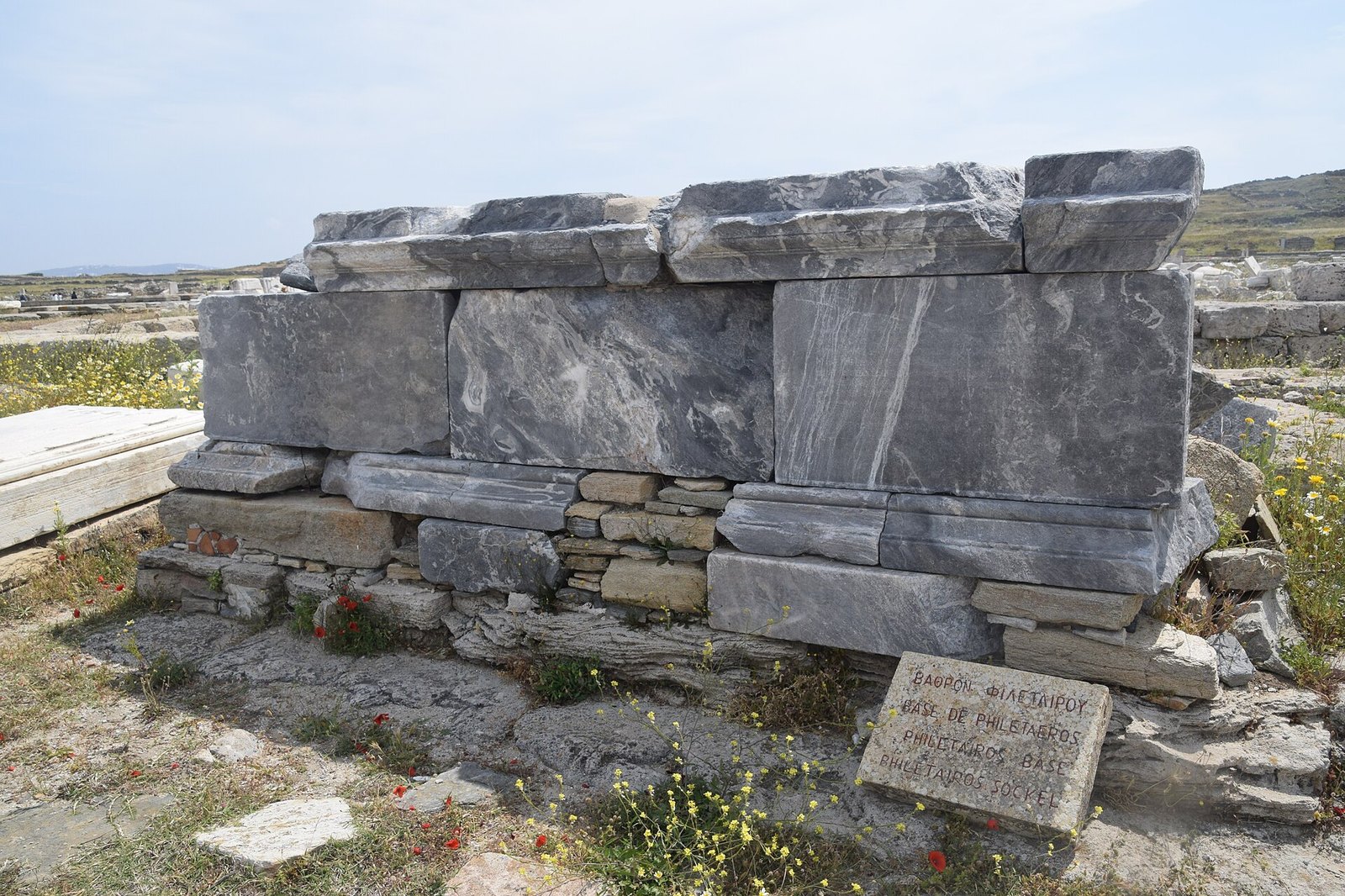 Large squared marble blocks stacked on a low stone wall at the Delos archaeological site, with grass and wildflowers around them