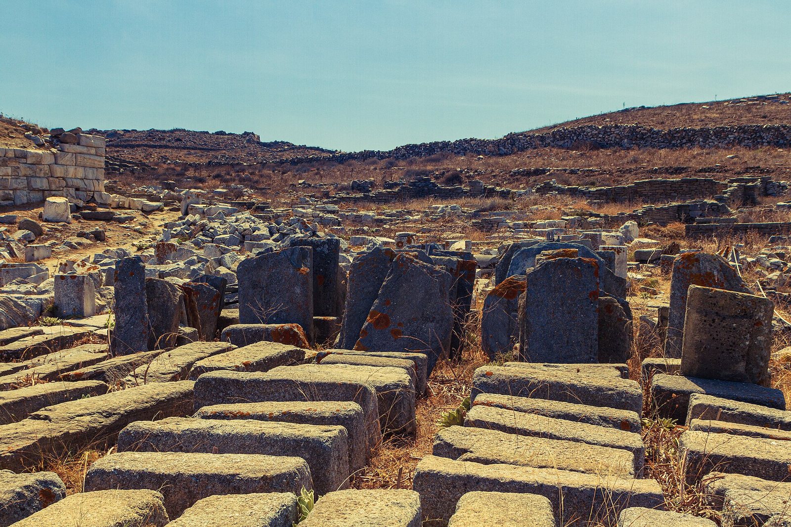 Numerous large, cut stone blocks and fragments are scattered across a dry, rocky archaeological site under a clear blue sky.