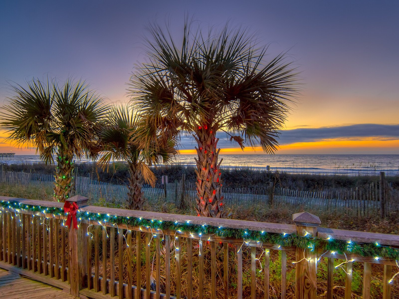 Palm trees strung with Christmas lights beside a wooden boardwalk railing, with the ocean and an orange sunset sky behind