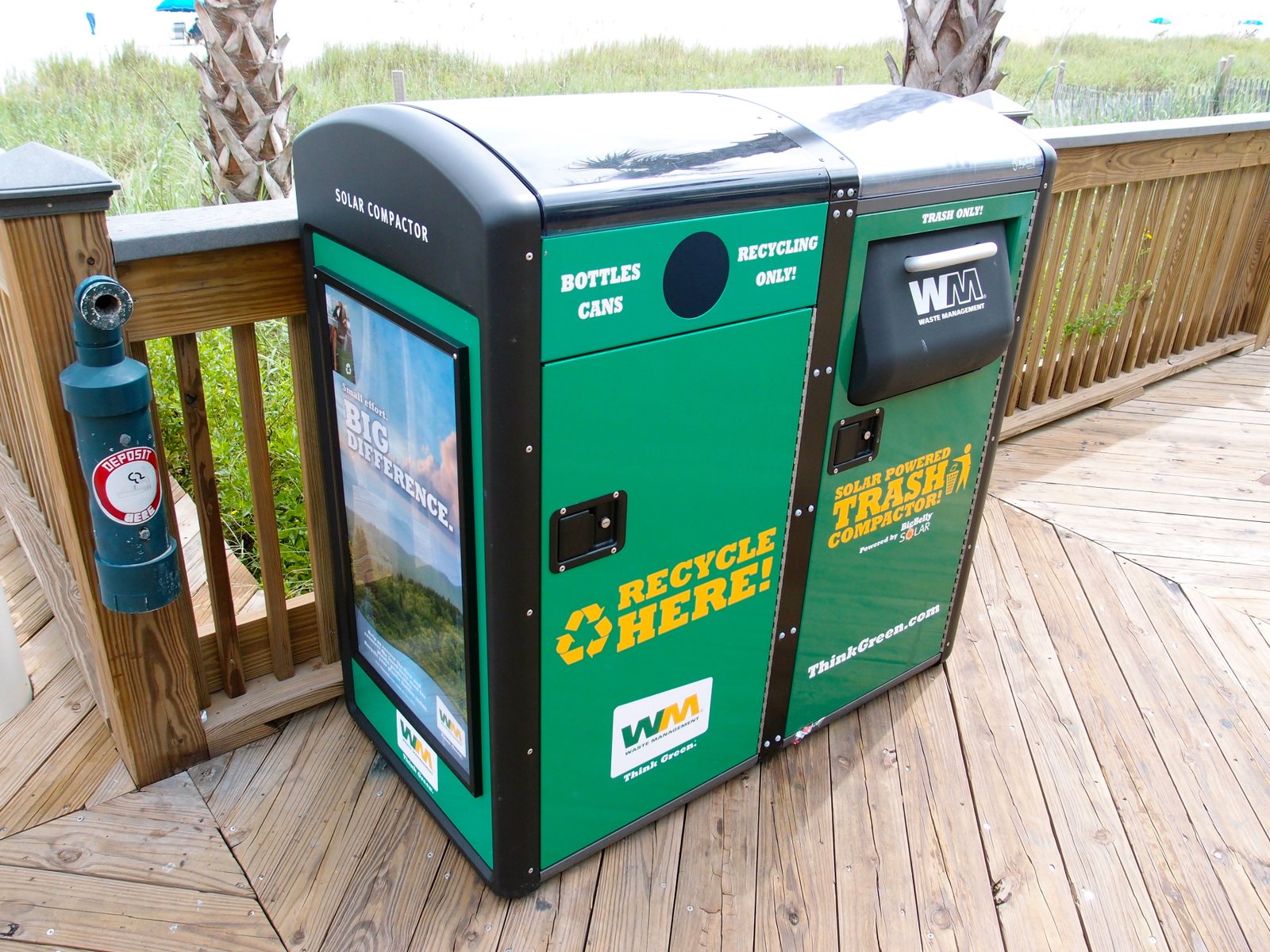 A green and black solar-powered recycling and trash compactor bin stands on a wooden boardwalk, with a blue deposit tube on a railing nearby.