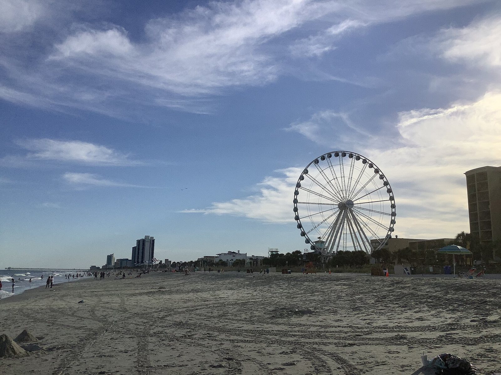 A wide sandy beach with people, a pier, high-rise buildings, and the SkyWheel Ferris wheel under a cloudy sky.