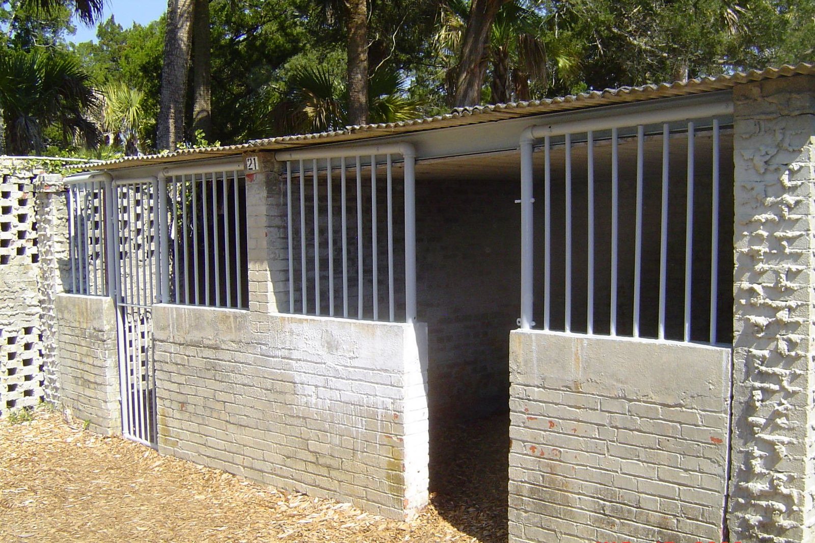 A row of brick and concrete horse stalls with metal bars and a corrugated roof stands on wood chip ground, with trees in the background.