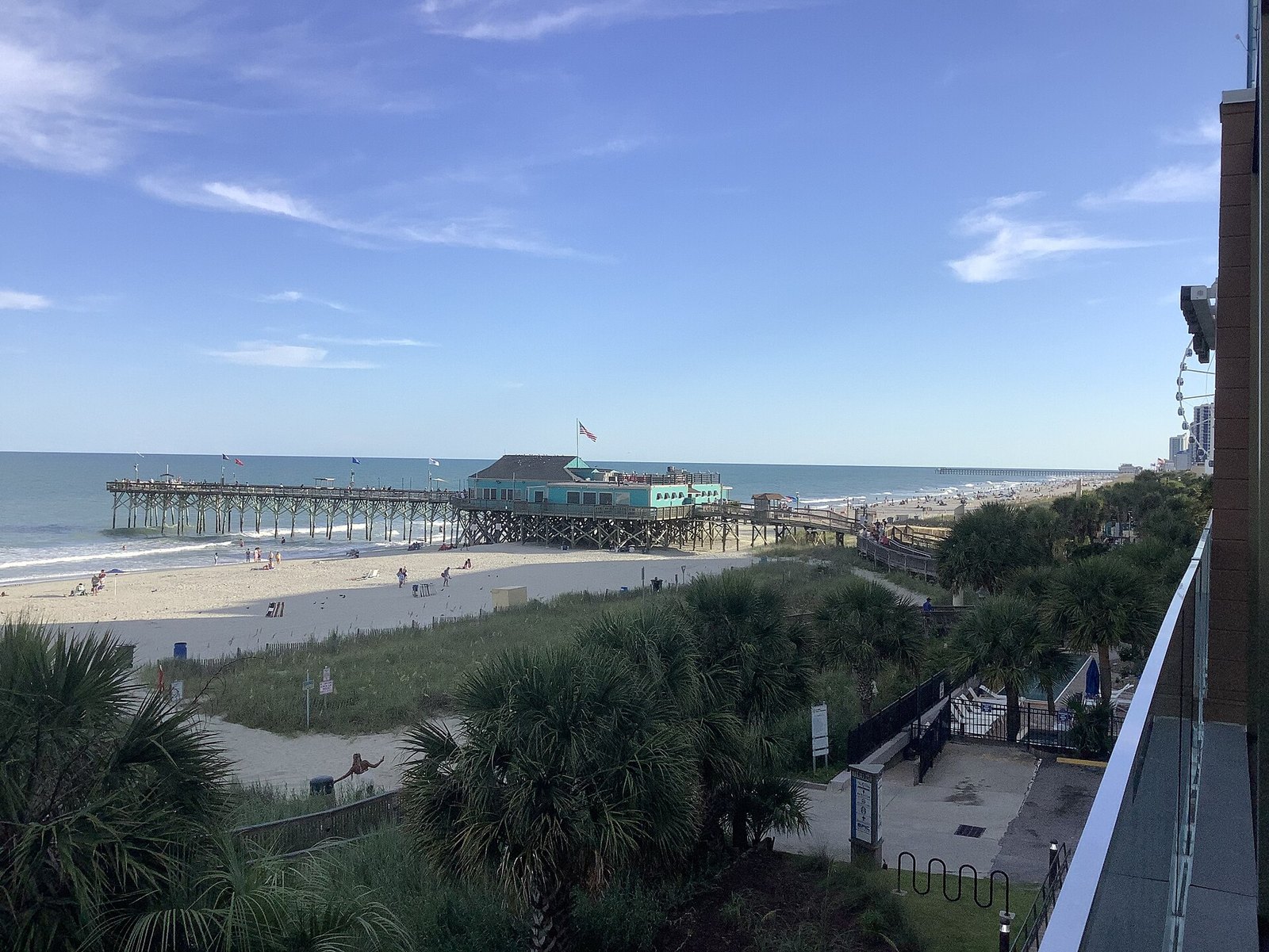 A long pier with a pavilion at its end extending into the ocean, seen from a balcony above dune grass and a sandy beach under a blue sky