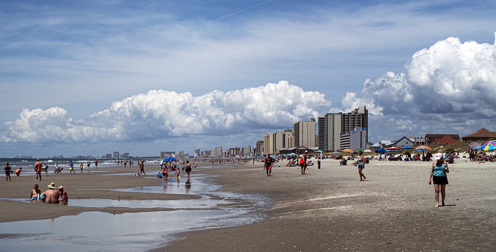 A wide sandy beach is busy with people walking and relaxing, with high-rise buildings and houses visible along the distant coastline under a blue sky with large white clouds.