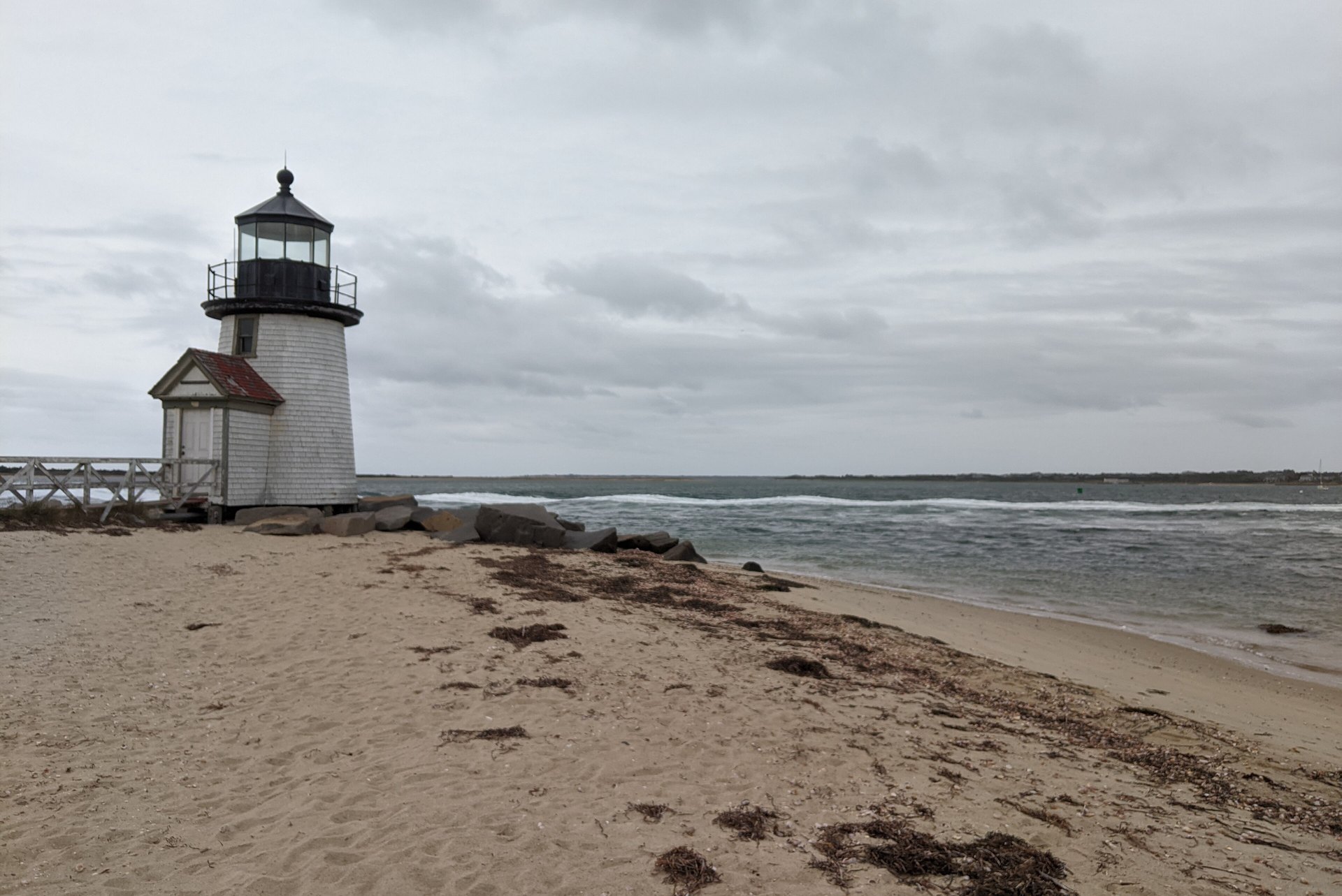 The short white Brant Point Light with its red roof standing at the sandy entrance to Nantucket harbour, with grey surf and an overcast sky