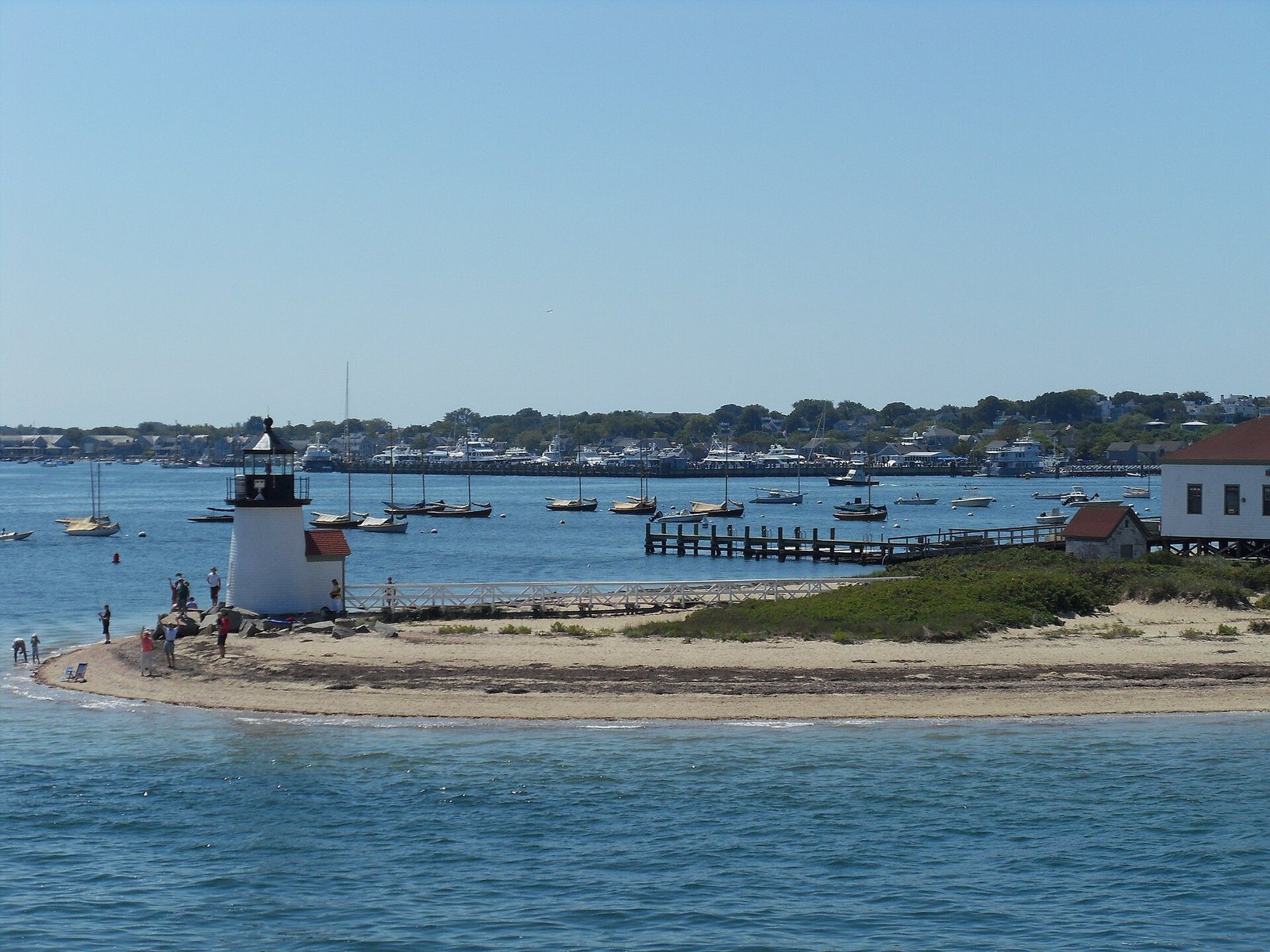 A white lighthouse with a black top stands on a sandy point, with people nearby, overlooking a harbour filled with boats and a town in the background.