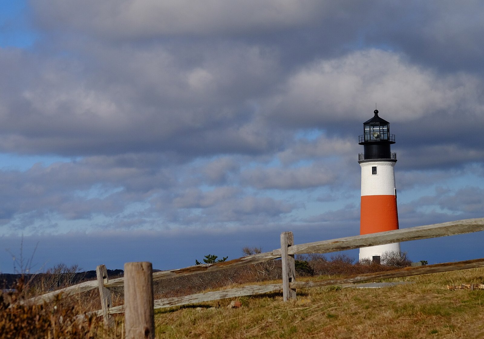 A red-banded white lighthouse with a black lantern on a grassy headland behind a split-rail fence, under dark clouds