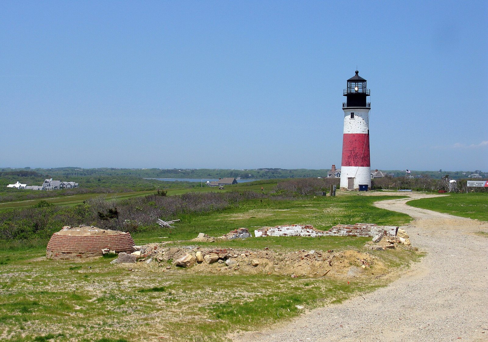 A red and white lighthouse stands on a grassy hill with a dirt path leading towards it, alongside some brick ruins and distant houses under a clear blue sky.