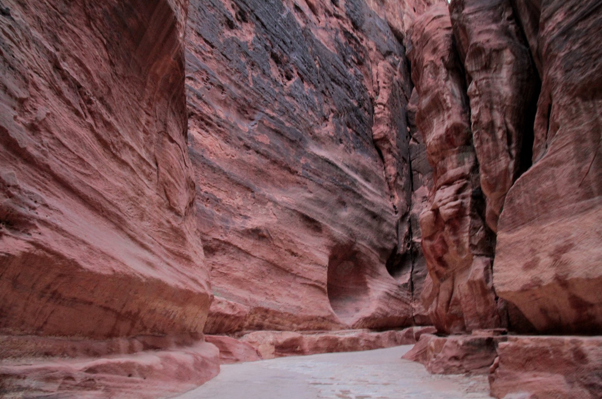 A narrow, high-walled sandstone gorge with a pale floor curving between towering red and pink striped rock faces