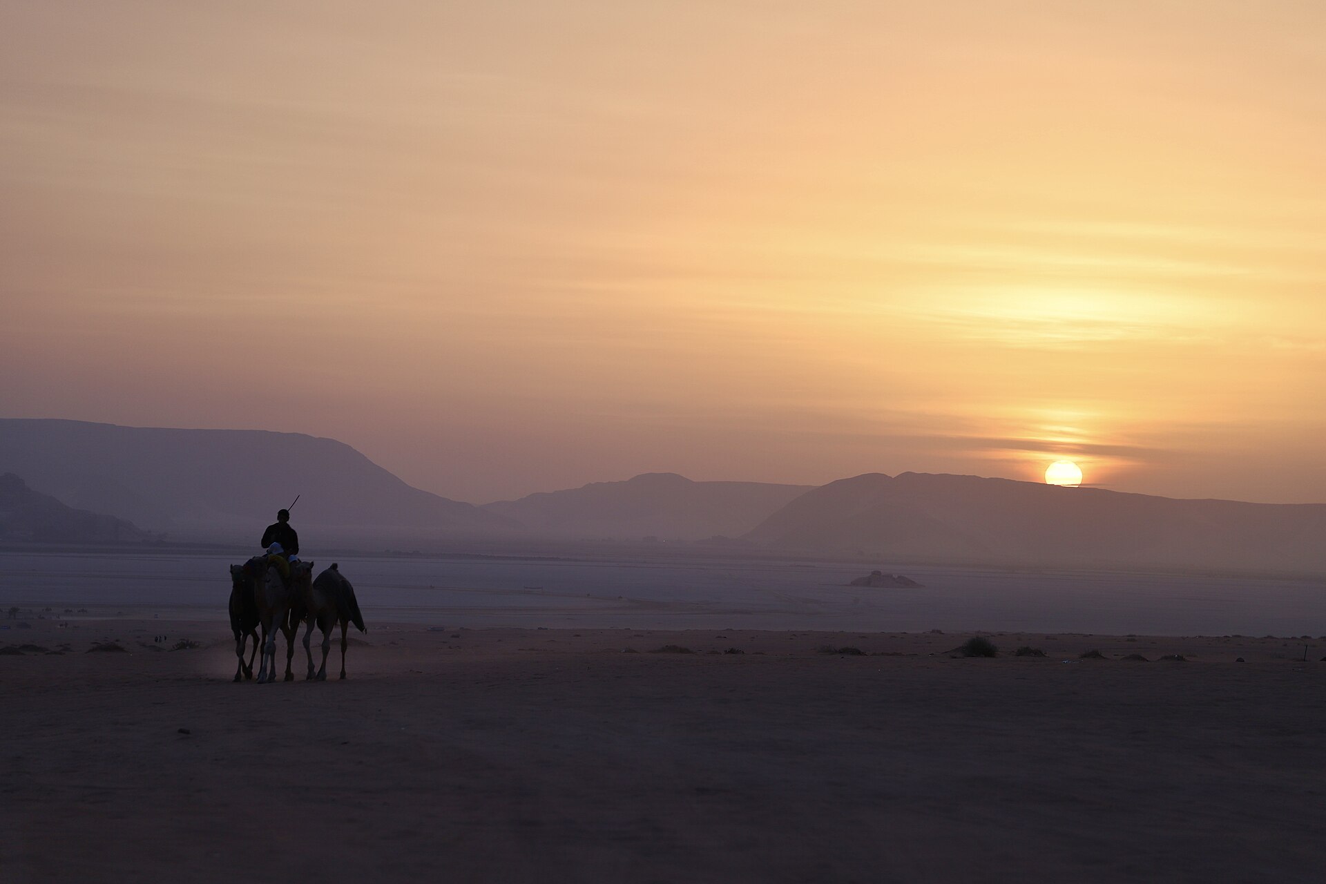A wide red-sand desert valley with steep sandstone and granite mountains rising from the flat floor under a clear sky.