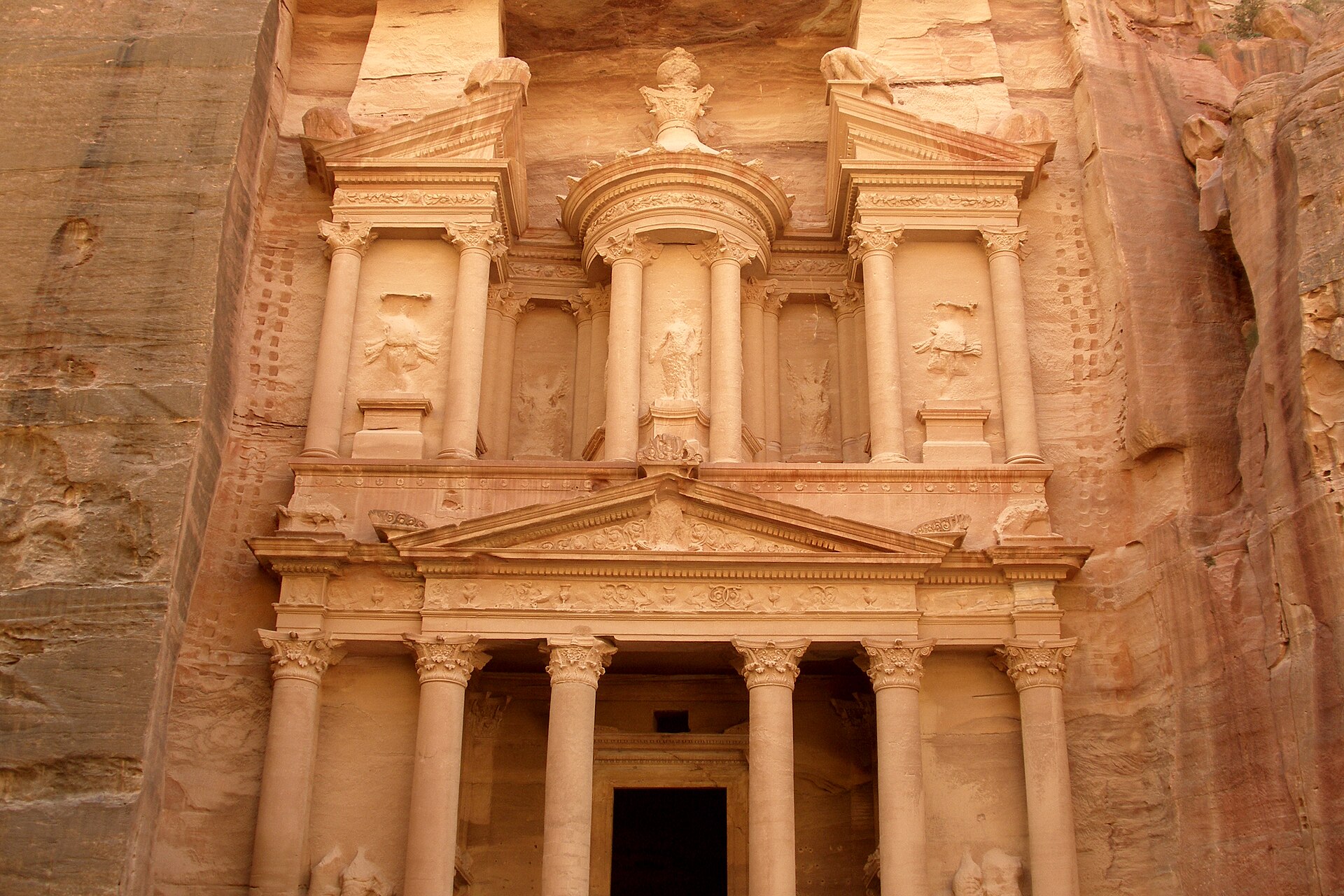 A tall carved sandstone façade with columns and an urn, framed by the walls of a narrow rock gorge in warm morning light.