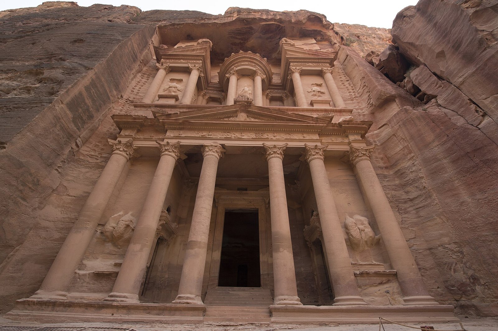 The carved sandstone facade of the Treasury, with tall columns and a pediment, seen from below in a rose-red gorge