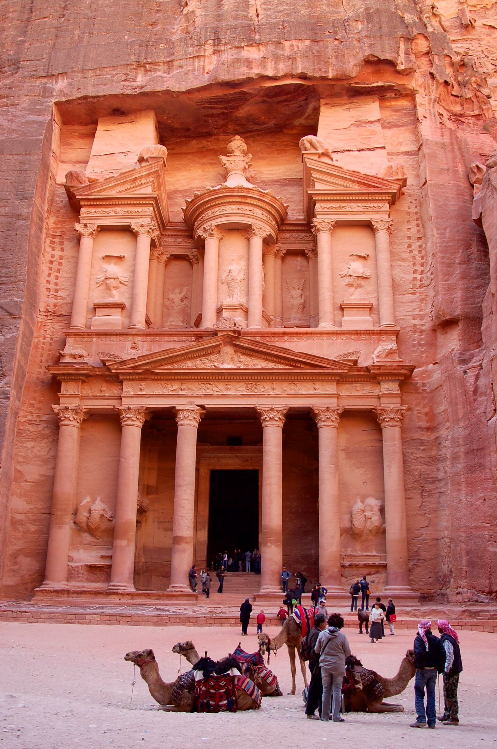 Two decorated camels rest on sandy ground before the rock-carved facade of Khaznat el- Faroun or Pharaoh's treasury, with people nearby.
