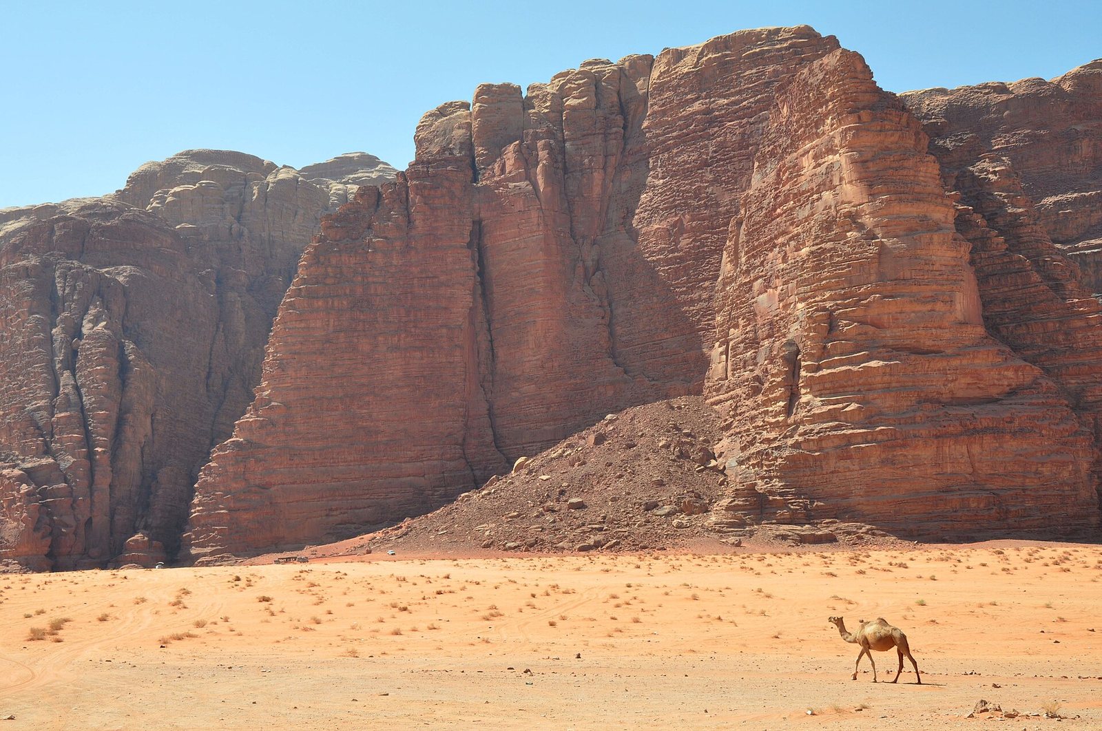 A carved sandstone monument, Al-Khazneh (Treasury façade), is seen from above, with three people on a rocky ledge.
