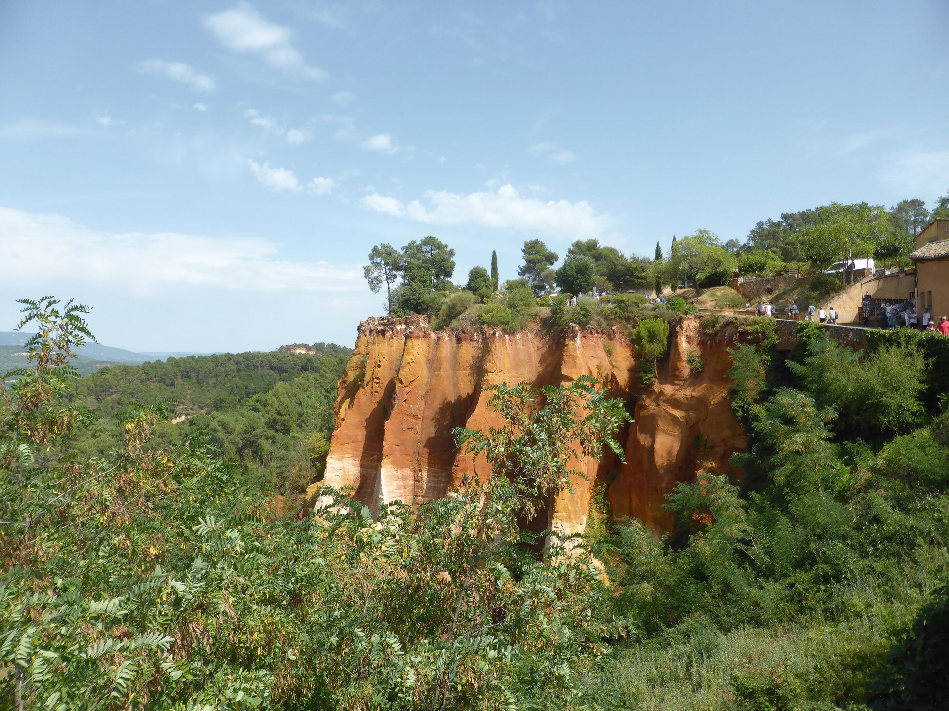 A cliff of orange and red ochre rock above green woodland at the start of the ochre trail in Roussillon, under a blue sky