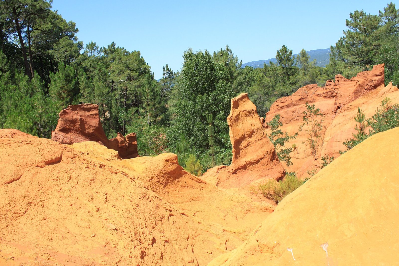 A landscape view of orange and reddish-brown ochre rock formations with green trees and shrubs under a clear blue sky.