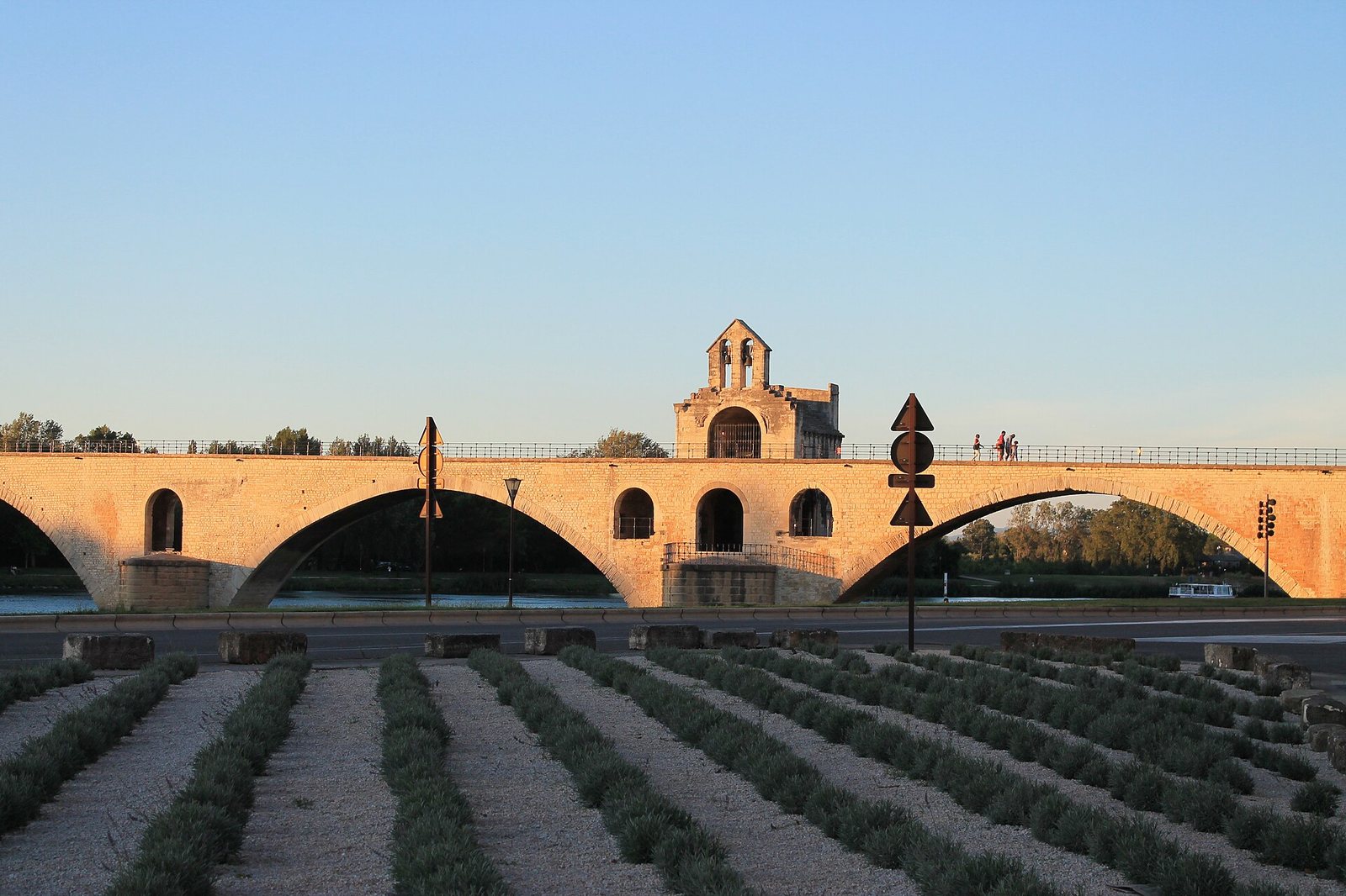 A stone bridge with arches and a chapel-like structure spans a river, with rows of low green plants in the foreground under a clear sky.
