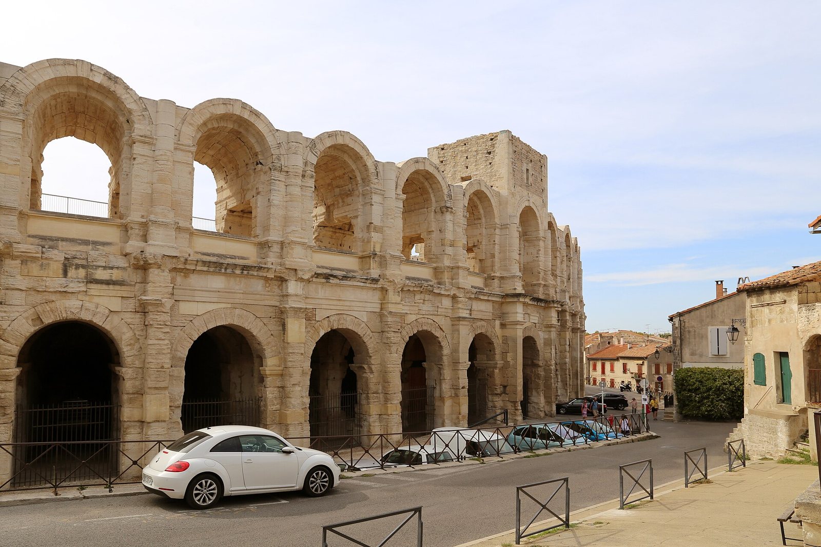 The exterior of a large Roman amphitheater with multiple arched levels, a street with parked cars and pedestrians, and old buildings under a partly cloudy sky.