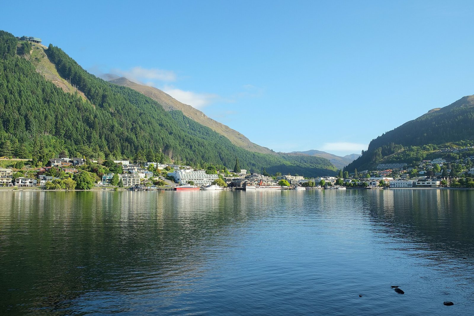 A wide view of Queenstown Bay with a town along the waterfront, surrounded by tree-covered mountains, under a clear sky. A gondola station is visible on the upper left mountain.