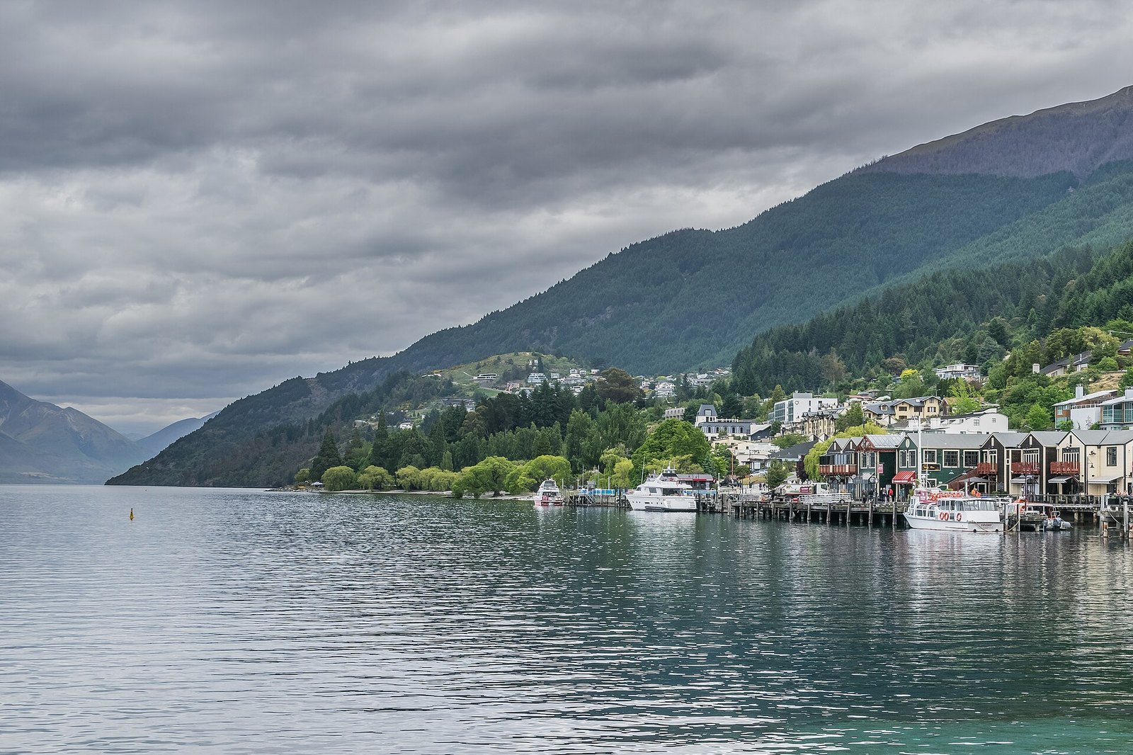 A calm lake beneath forested mountains under heavy cloud, with the waterfront of Queenstown and moored boats at the right