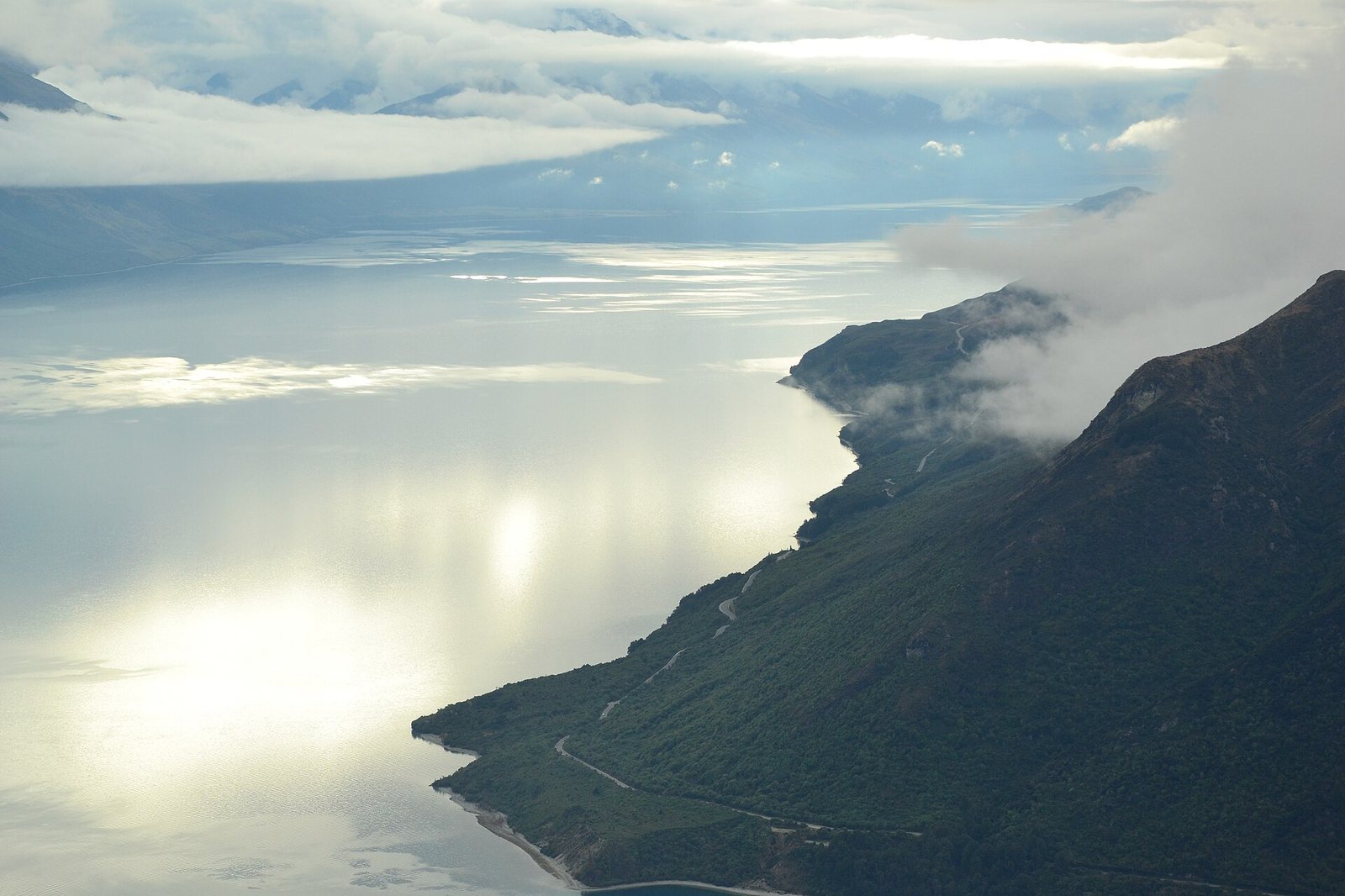 An aerial view shows a large lake reflecting sunlight, bordered by a forested mountain with a winding road, under a sky with clouds.